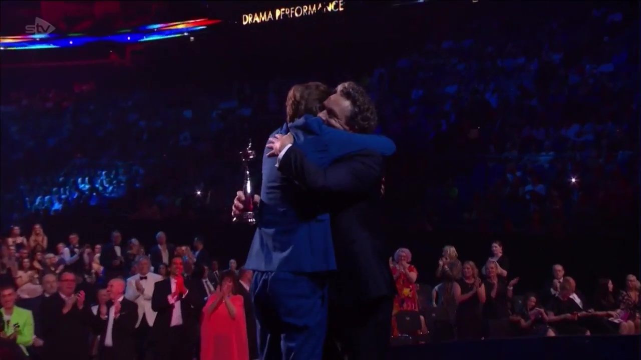 David Tennant and Michael Sheen hugging on stage while an audience stands in ovation.