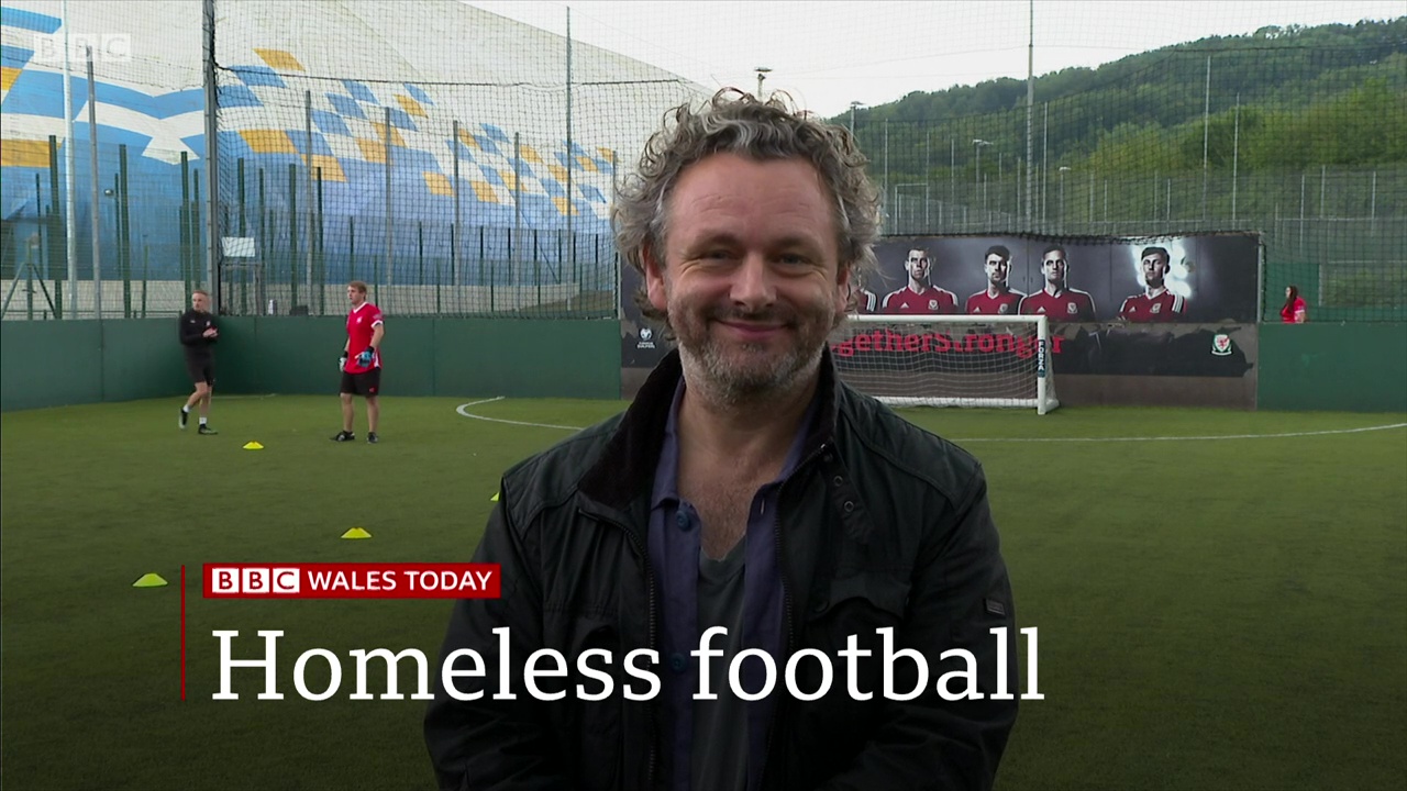 A still from Michael Sheen's appearance on BBC Wales Today. He stands on a football pitch with people playing behind him, he's wearing a black coat and a blue shirt, and has curly hair and a beard and is smiling. Text reads BBC Wales Today, Homeless football.