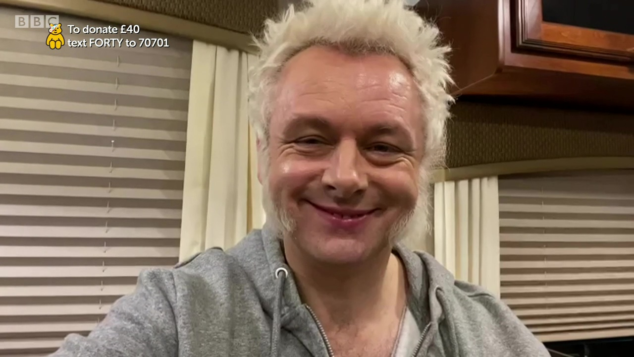 Michael Sheen in his trailer on the Good Omens set, wearing a grey hoody, with white sideburns and white hair, smiling.