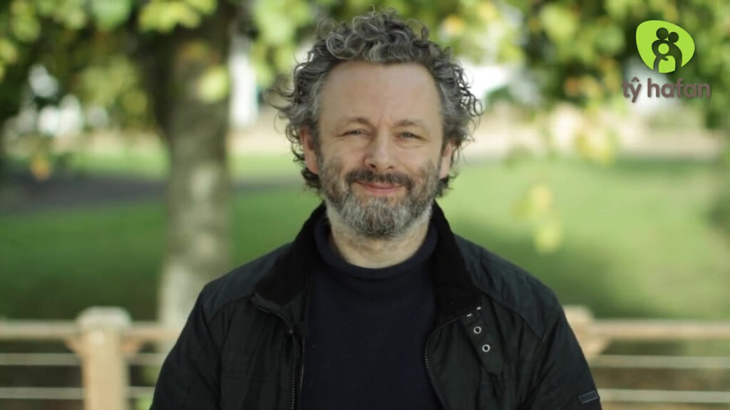 Michael Sheen with very curly hair and a beard, wearing a dark blue jumper and black coat, stands outside in front of trees and grass, smiling.