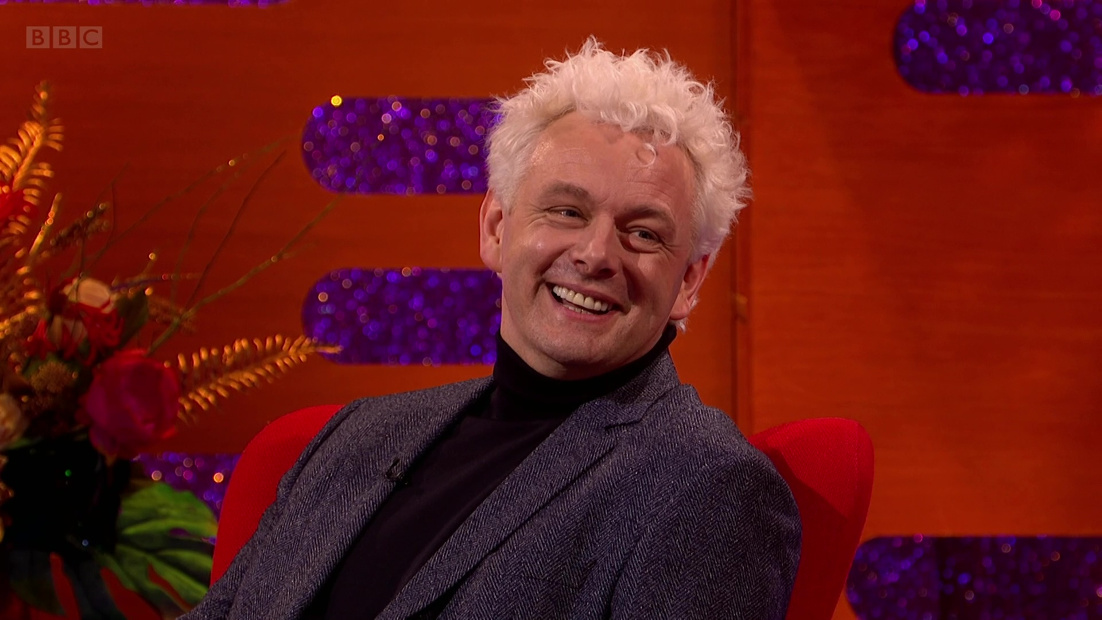 Michael Sheen with his white Aziraphale hair, wearing a black roll-neck jumper and a grey suit jacket, sitting in a red chair laughing, on the red and purple glittery Graham Norton set.