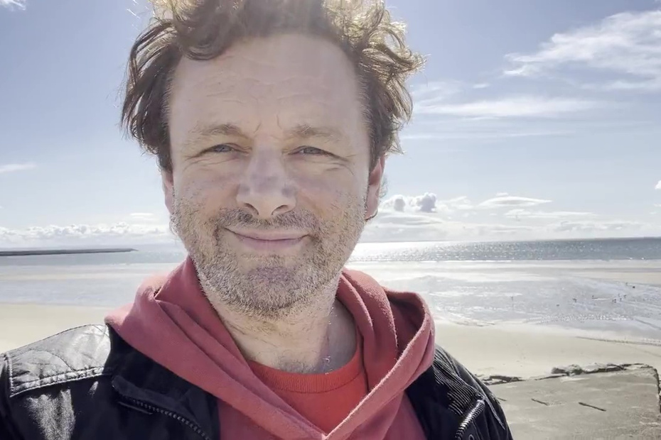 Michael Sheen at a beach with blue sky, wearing a red hoodie under a black jacket, smiling