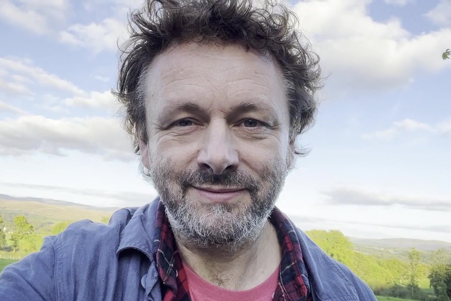 Michael Sheen in a blue jacket and red shirt outdoors in front of a blue sky with white clouds and green hills