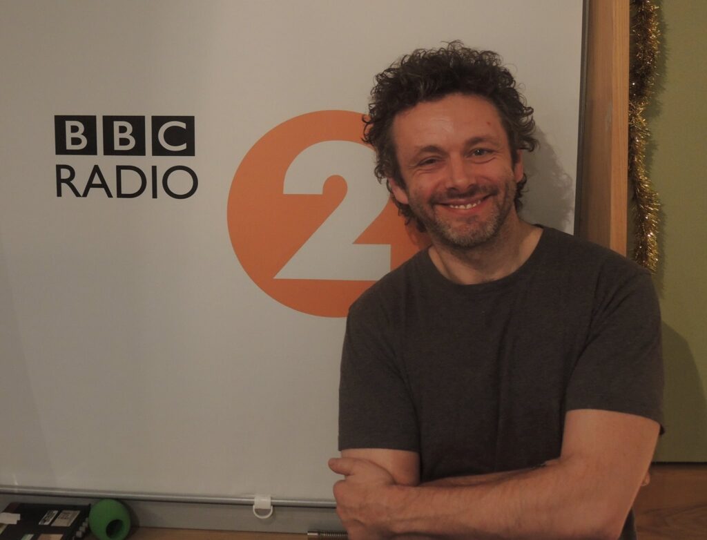 Michael Sheen standing in front of the BBC Radio 2 logo.