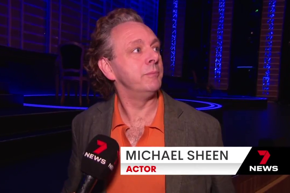 Michael Sheen in a grey suit jacket with an orange shirt, beardless with curly grey hair, being interviewed for the news in the Sydney Opera House.