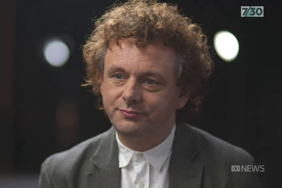 Michael Sheen clean shaven in a grey suit jacket and white shirt.